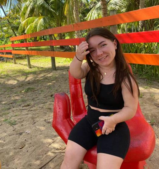 Girl sitting on a red chair shaped like a hand in front of an orange fence and palm trees.